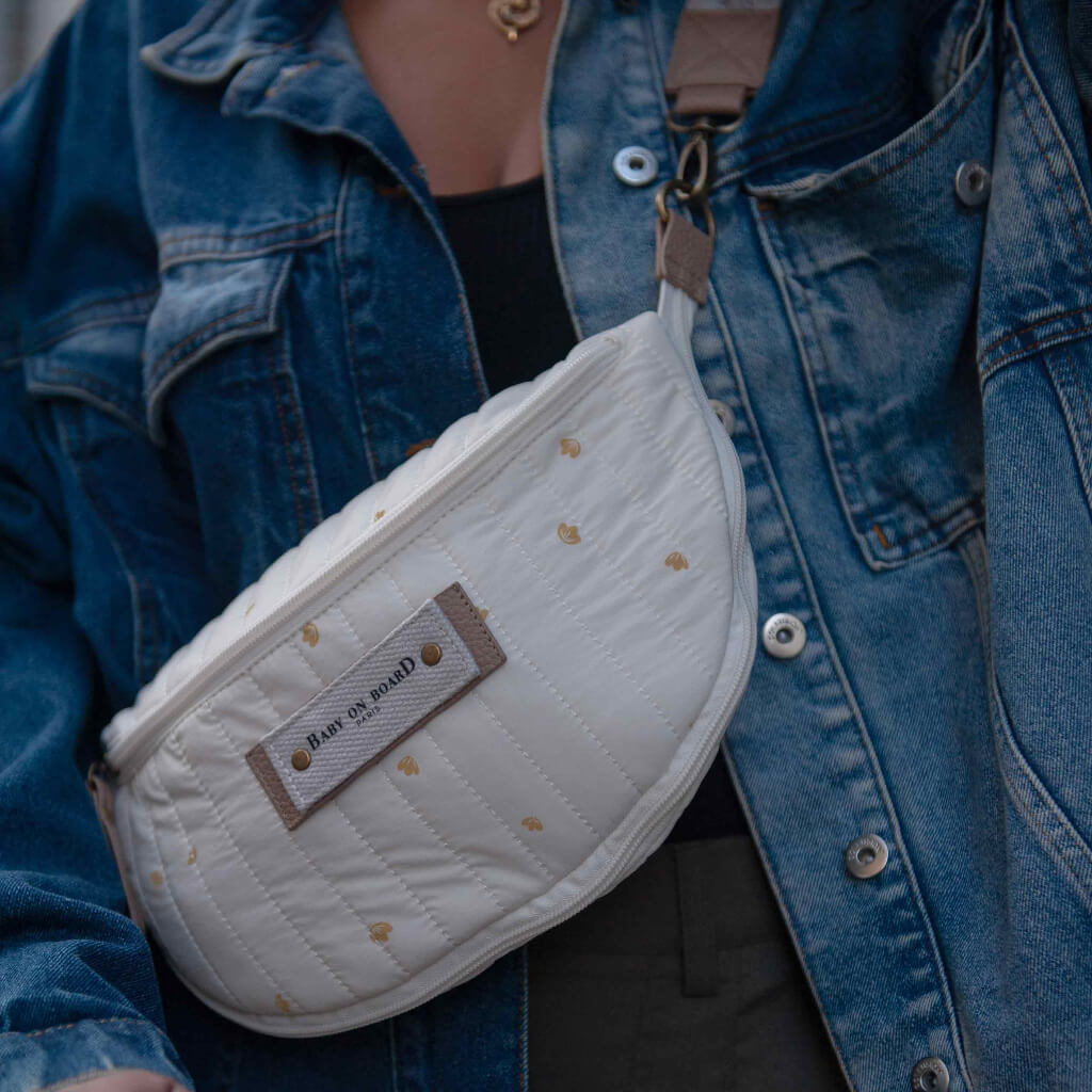 Sac banane à langer Armelle blanc crème Baby on Board porté en bandoulière sur une veste en jean, un accessoire tendance et pratique pour les parents.